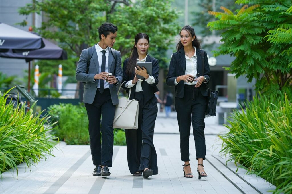 Three professionals walking along a landscaped outdoor path, carrying a tablet and takeaway cups